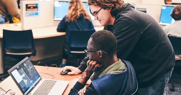 Two students in an ETSU computer lab. One student sits a desk while the other is standing above him, pointing to something on the computer screen.