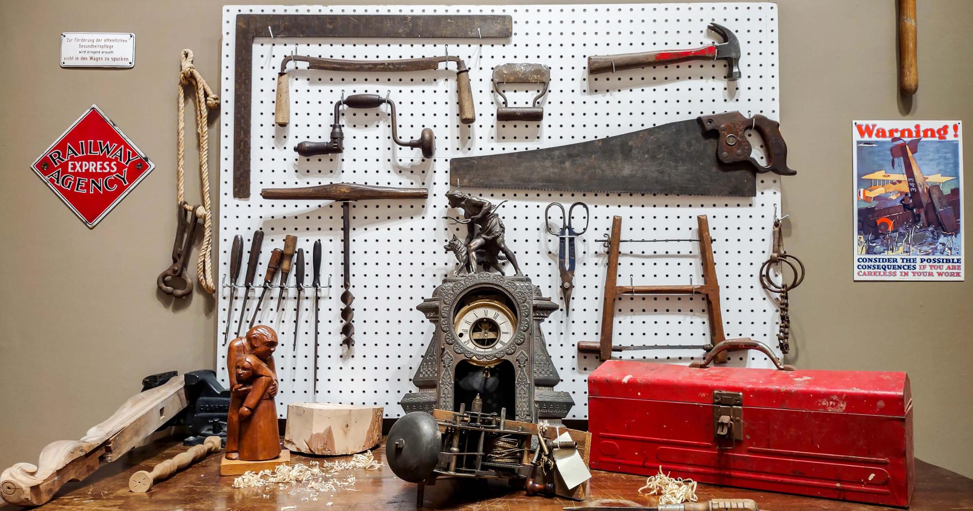 Image depicts tools held on a pegboard. A table with wood working artifacts sits below.