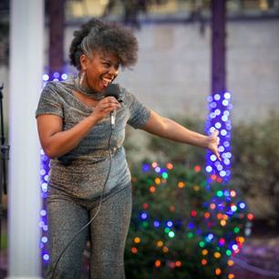 Woman standing on a stage speaking into a microphone with Christmas lights in the background