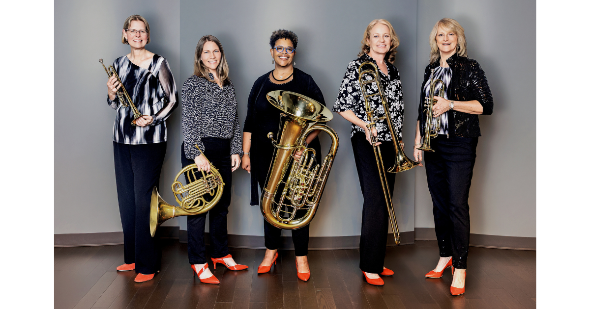 Five women brass musicians pose against a gray studio backdrop, holding their instruments: two trumpets, a French horn, a tuba, and a trombone. They wear coordinated black outfits with patterned tops and bright red-orange shoes. The group smiles at the camera, standing on a dark wood floor.