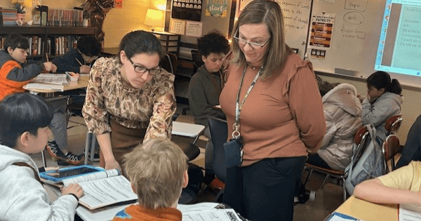 Two women standing over 2 students sitting at desks in elementary classroom. 