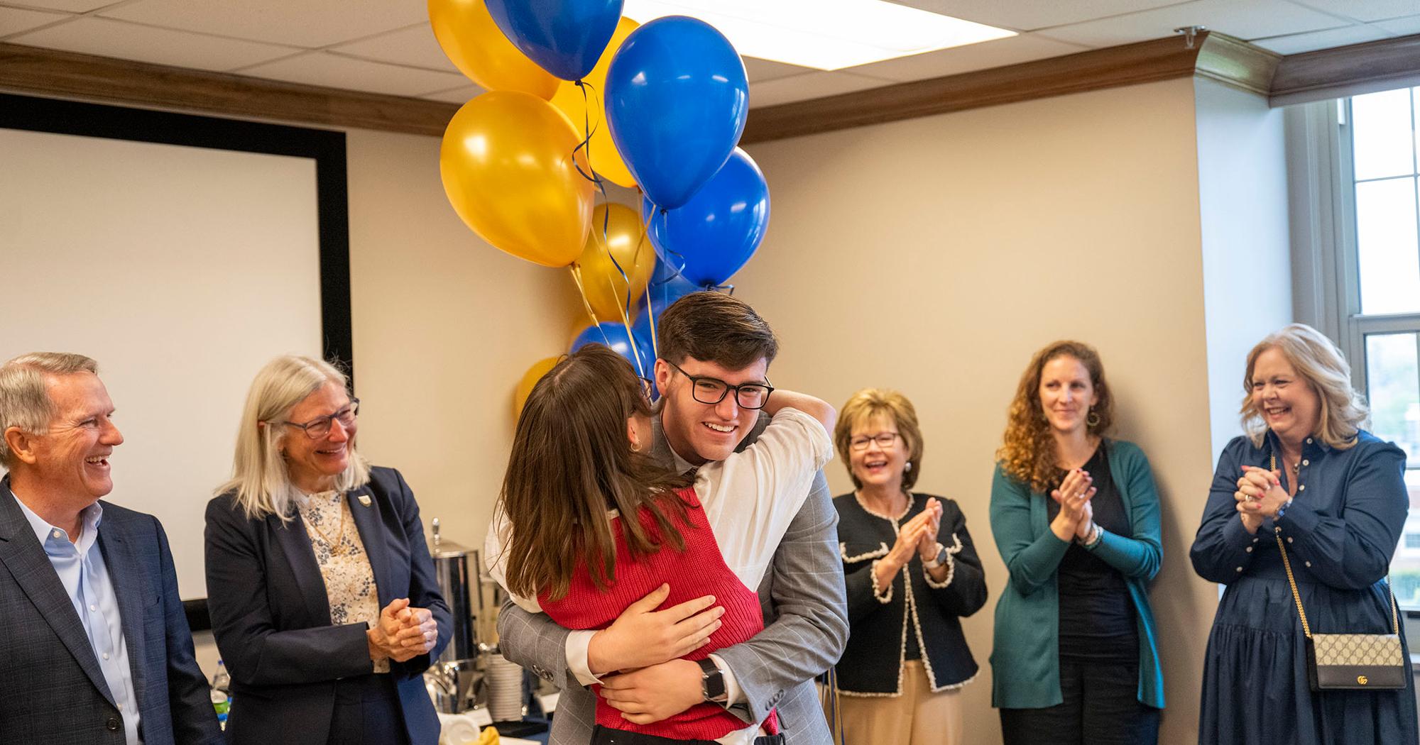 Student celebrates with a hug and smile during a surprise Truman Scholarship announcement at ETSU, surrounded by applauding faculty and staff with gold and blue balloons in the background.