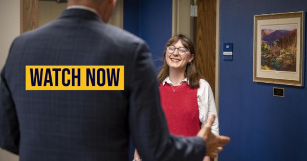 A smiling young woman in a red vest talks with a man in a suit, with the words "WATCH NOW" overlaid on the image. 
