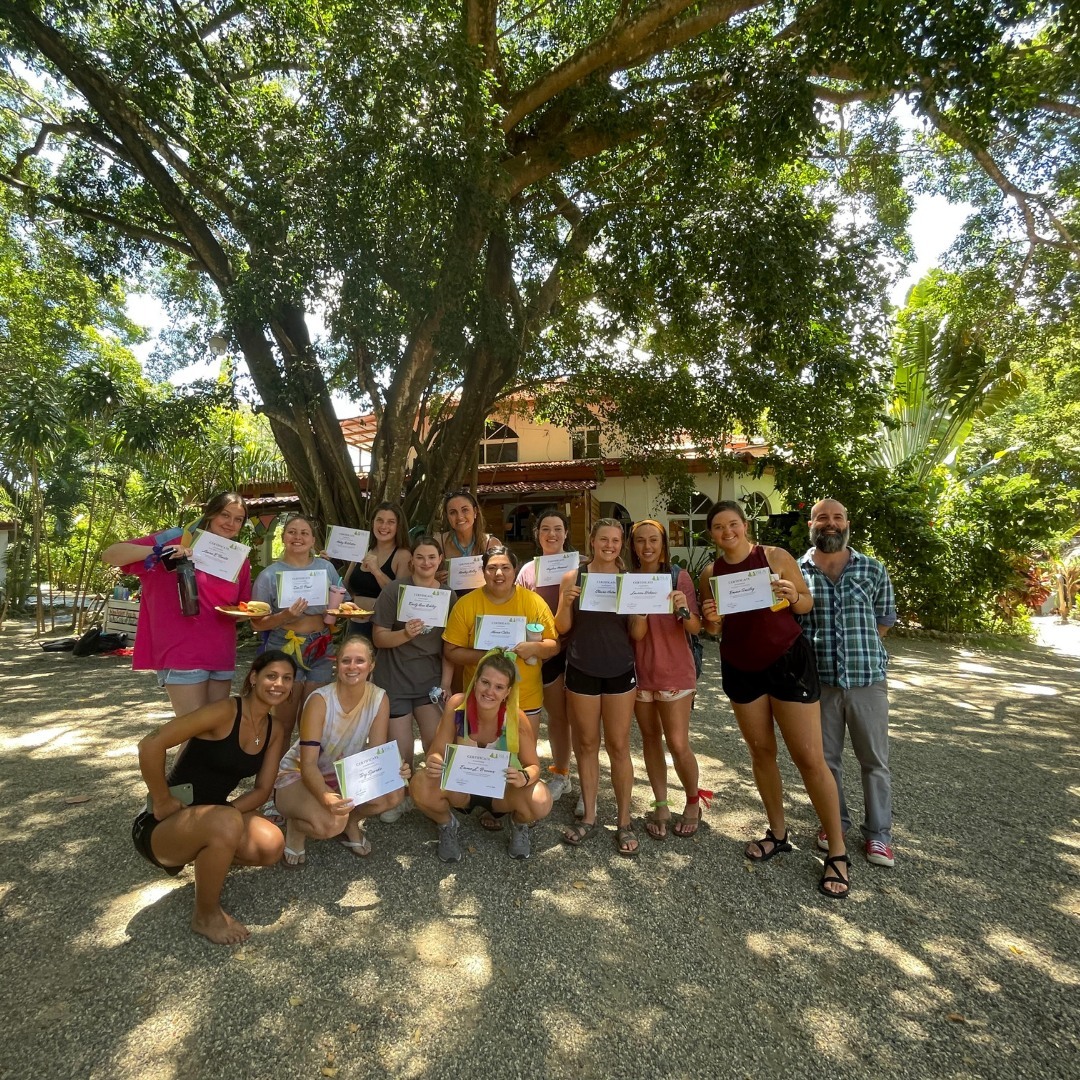 Students standing under a tree on the sand holding certificates