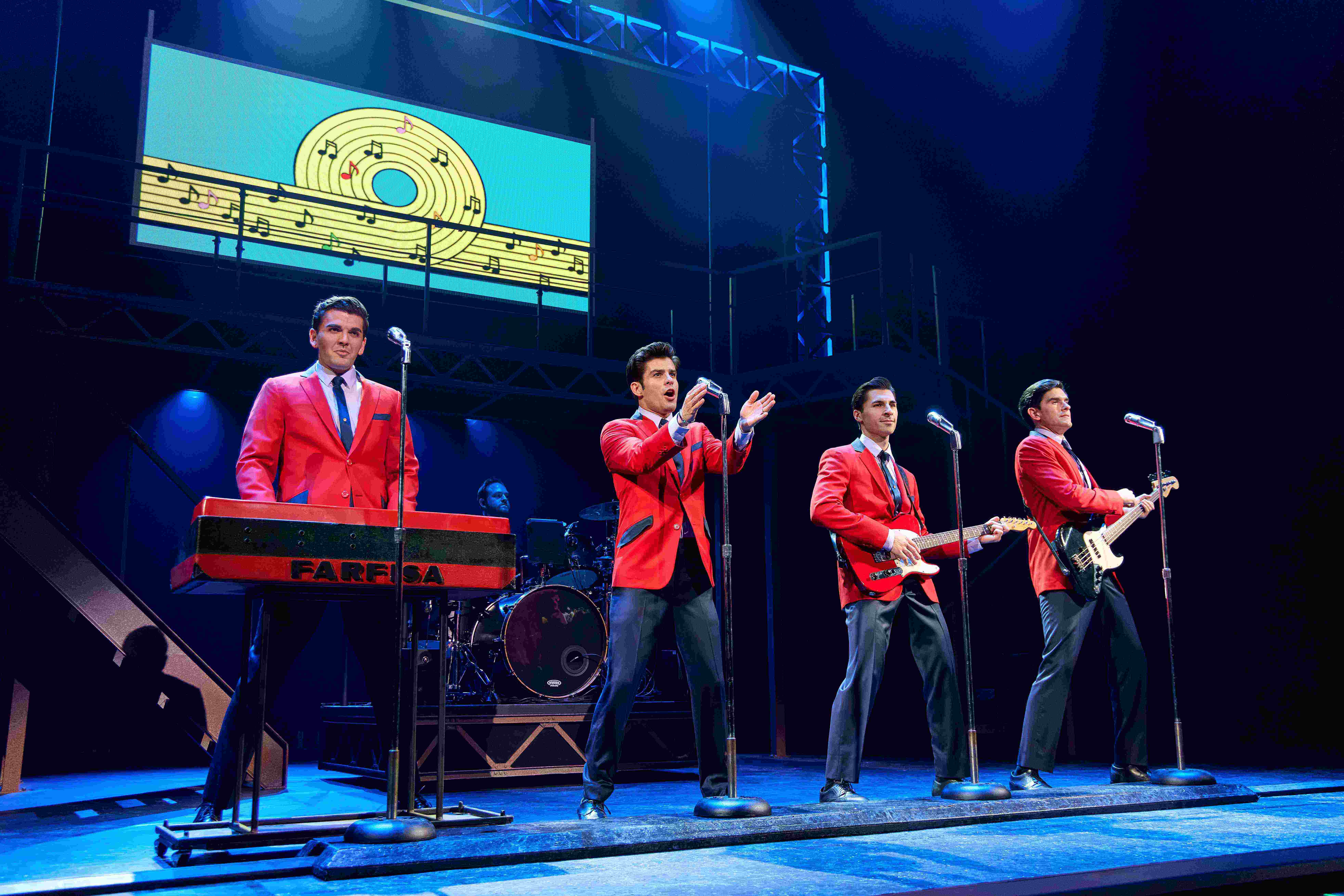 Stage photo from Jersey Boys showing four actors in matching red blazers and black trousers singing into microphones with raised arms.