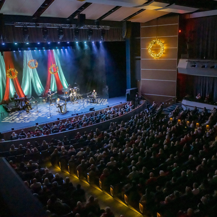High-angle shot of a live band performing on stage in a packed auditorium, illuminated by blue spotlights. Holiday wreaths decorate the background and walls.