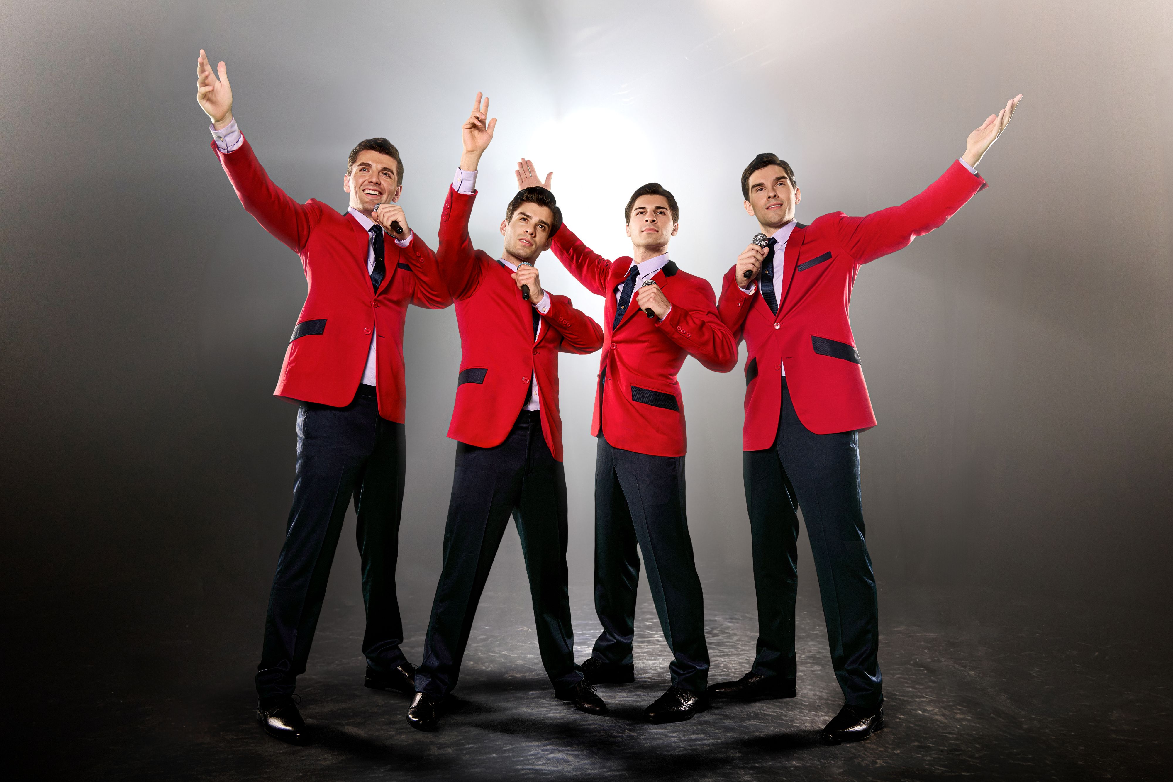 Stage photo from Jersey Boys showing four actors in matching red blazers and black trousers singing into microphones with raised arms.