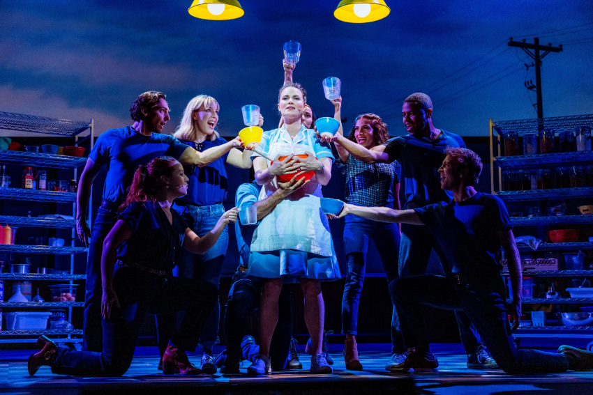 Stage photo from Waitress showing a woman in a pie diner uniform holding a red bowl, surrounded by cast members offering mixing cups and bowls.