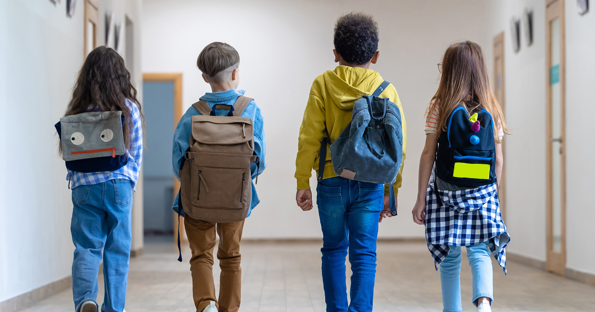 four young children, age 5 to 7, with backpacks walk in a school hallway. Their backs are to the camera.