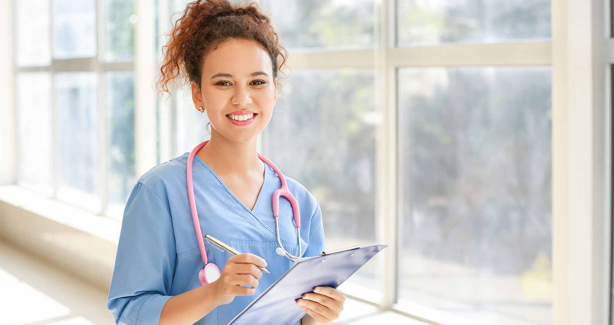 A young woman, a nursing students, stands in a healthcare facility with a stethescope and a clipboard.