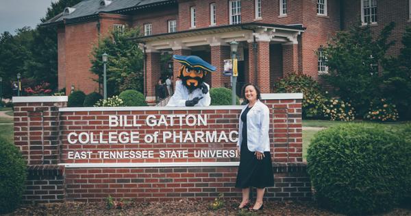 An ETSU pharmacy student standing outside on campus in front of the Bill Gatton College of Pharmacy sign. Bucky is hanging over the sign in a white coat and pointing to the student in excitement.