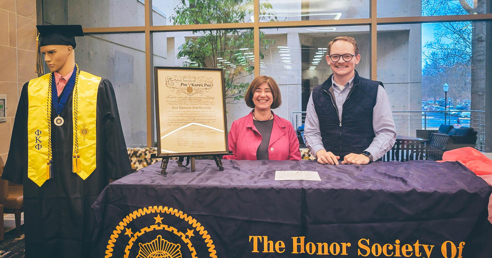 On the left, a mannequin adorned in Phi Kappa Phi regalia stands. The Phi Kappa Phi Honor Society table is visible, with two individuals standing behind the table smiling.