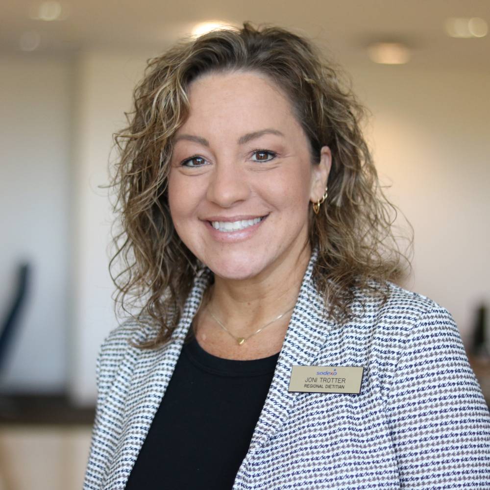 A photo of Joni Trotter, the ETSU Dietitian. She is wearing a blue and white blazer with a navy shirt. She has curly hair with highlights and she is smiling at the camera. 